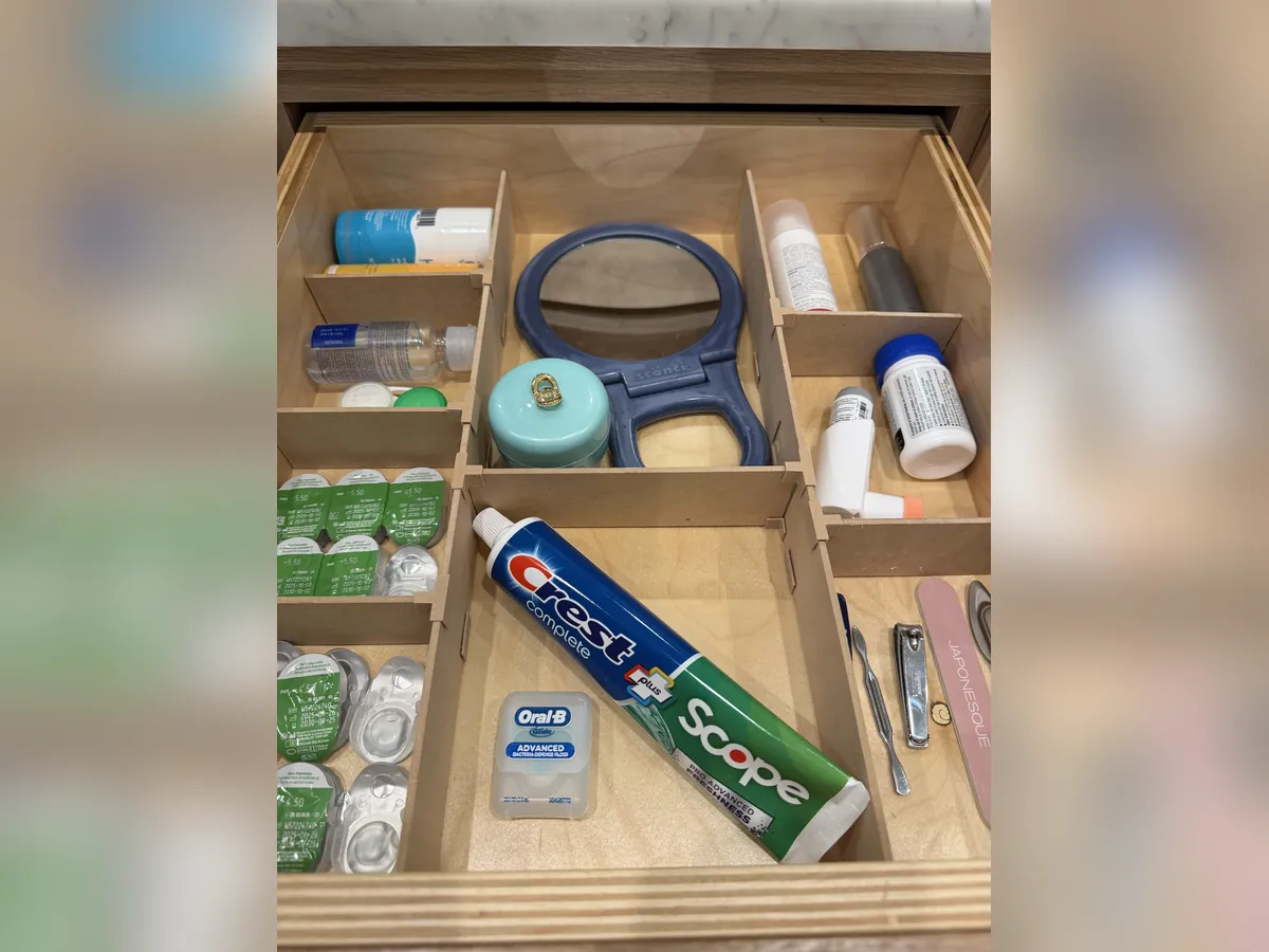 A bathroom drawer fitted with an Object Containment Device: a hand mirror, toothpaste and floss, contact lenses, and nail clippers and a file each in their own compartment