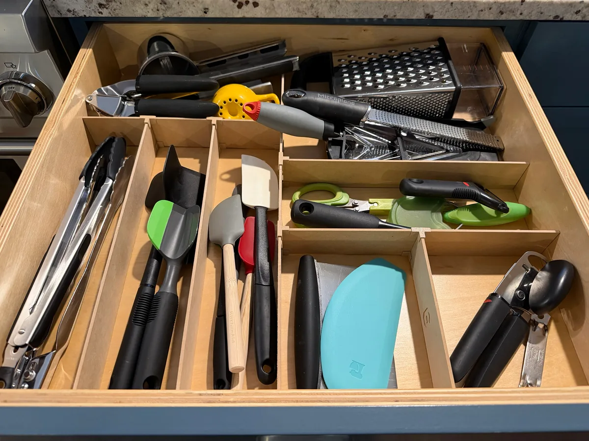 A kitchen drawer fitted with an Object Containment Device: tongs, spatulas and serving spoons standing in long slots on the left, graters and a citrus press at the back, and scissors, peelers, scrapers and a can opener in the compartments on the right